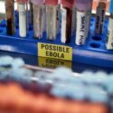 Et blåt reagensglasstativ indeholder flere blodprøverør; på en gul etiket på stativet står der "MULIG EBOLA". Scenen ser ud til at være fra et laboratorium.