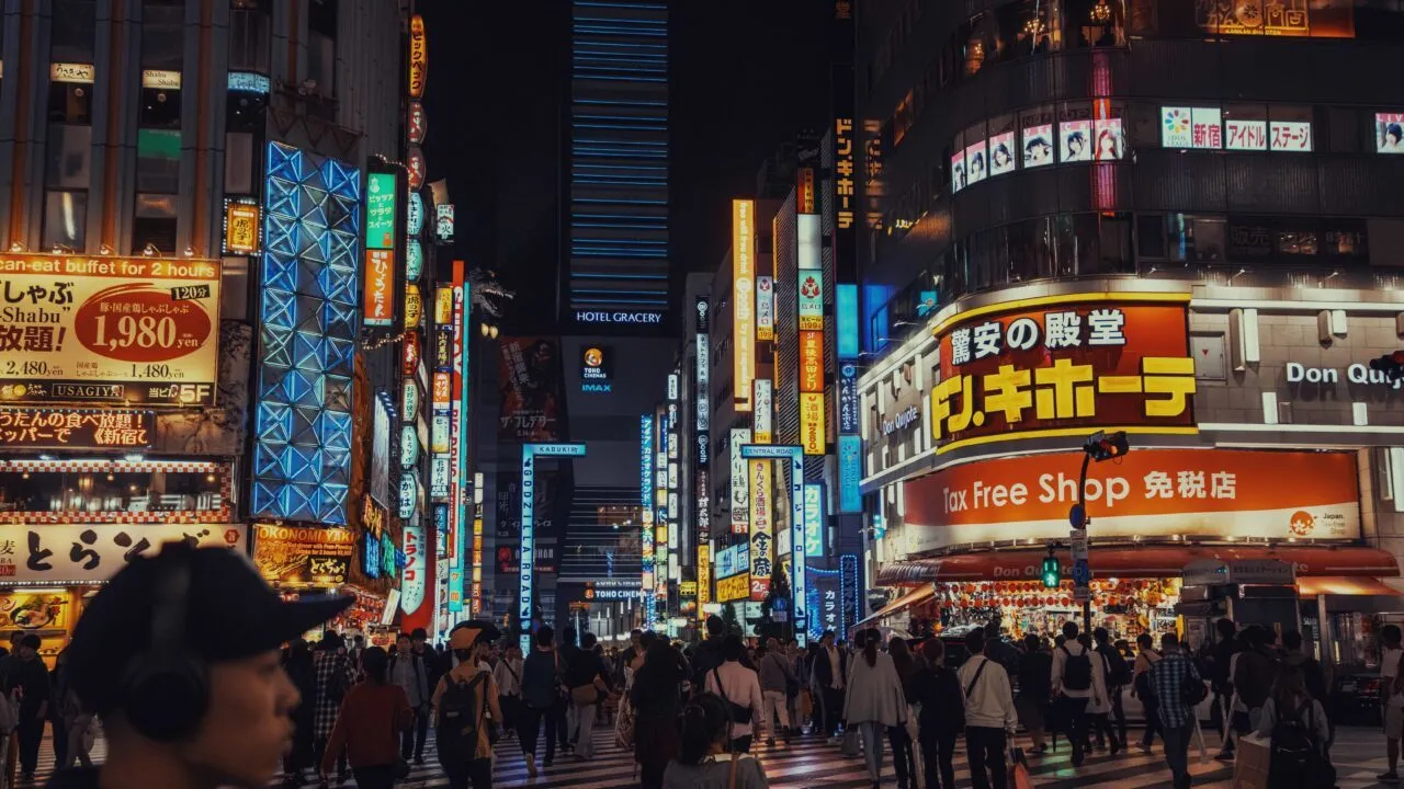 En travl gade i Tokyo om natten, fyldt med folk, der krydser vejen, omgivet af lyse, farverige neonskilte og høje bygninger, herunder hoteller og butikker, der lyser op i bybilledet.