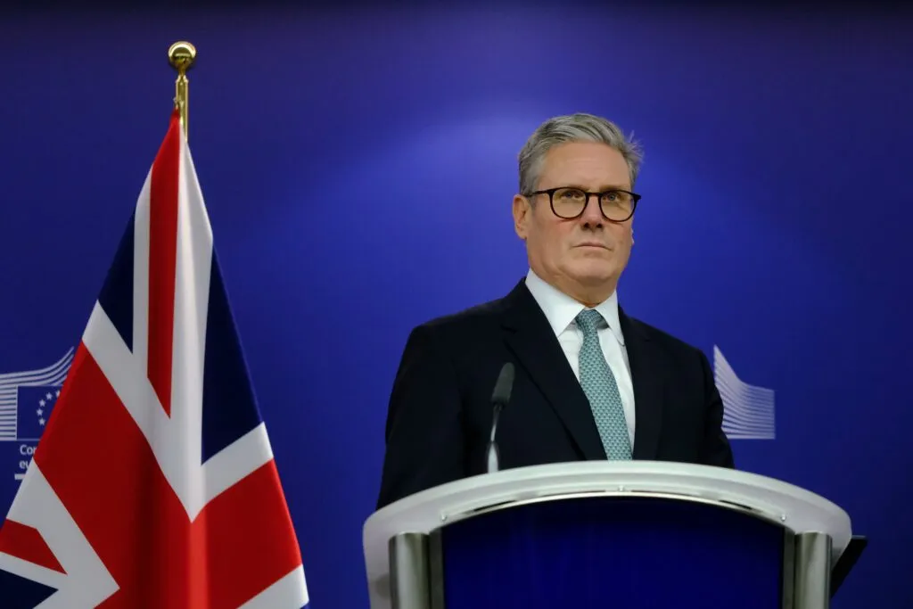 En mand i jakkesæt og briller står ved et talerstol med mikrofoner ved siden af et britisk flag foran en blå baggrund med EU-symboler.