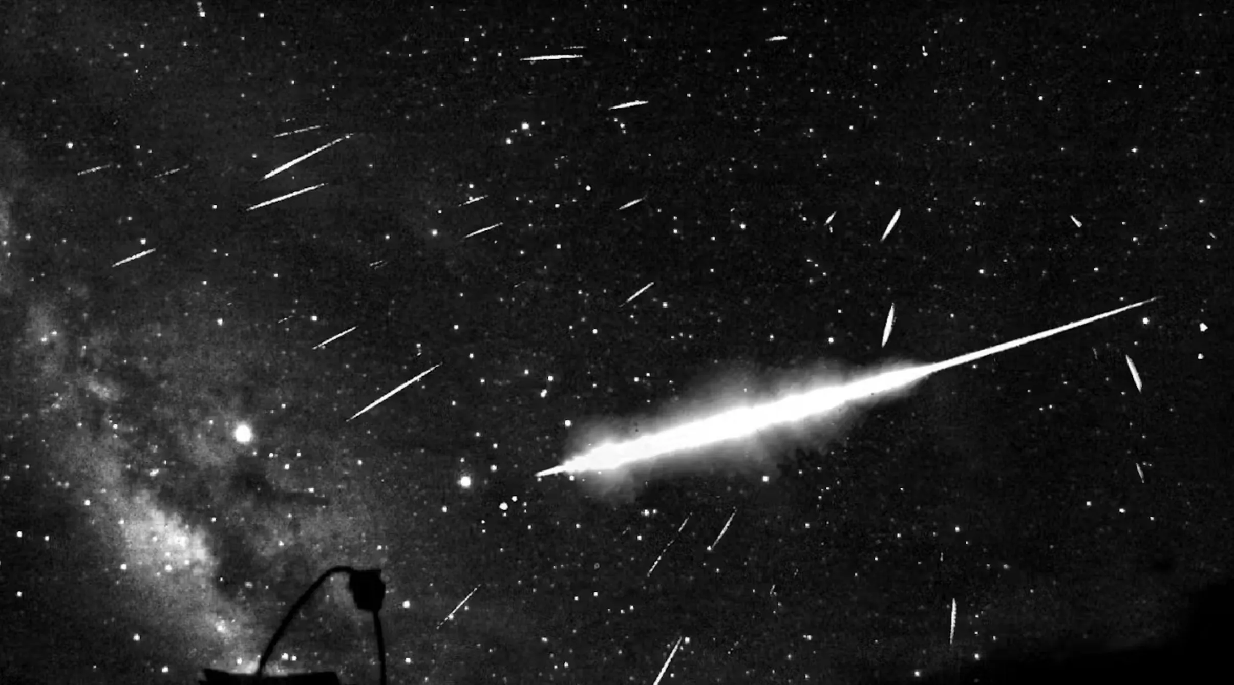 Et sort-hvidt foto, der viser en lysende meteor, der stryger hen over en stjernefyldt nattehimmel, med mange mindre meteorer synlige og en svag silhuet af udstyr eller en struktur i bunden.