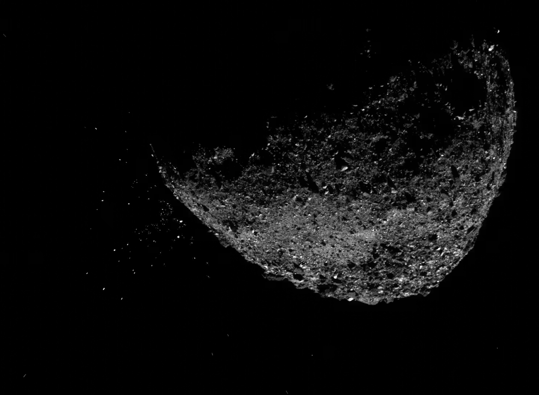 En stenet, halvt oplyst asteroide svæver i rummets mørke med små partikler og vragrester spredt rundt på overfladen mod en helt sort baggrund.