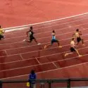 Seks løbere sprinter på en buet del af en udendørs bane, hver i deres egen bane. En person i blåt står ved rækværket og observerer løbet. Banens overflade er rødbrun med hvide banemarkeringer.