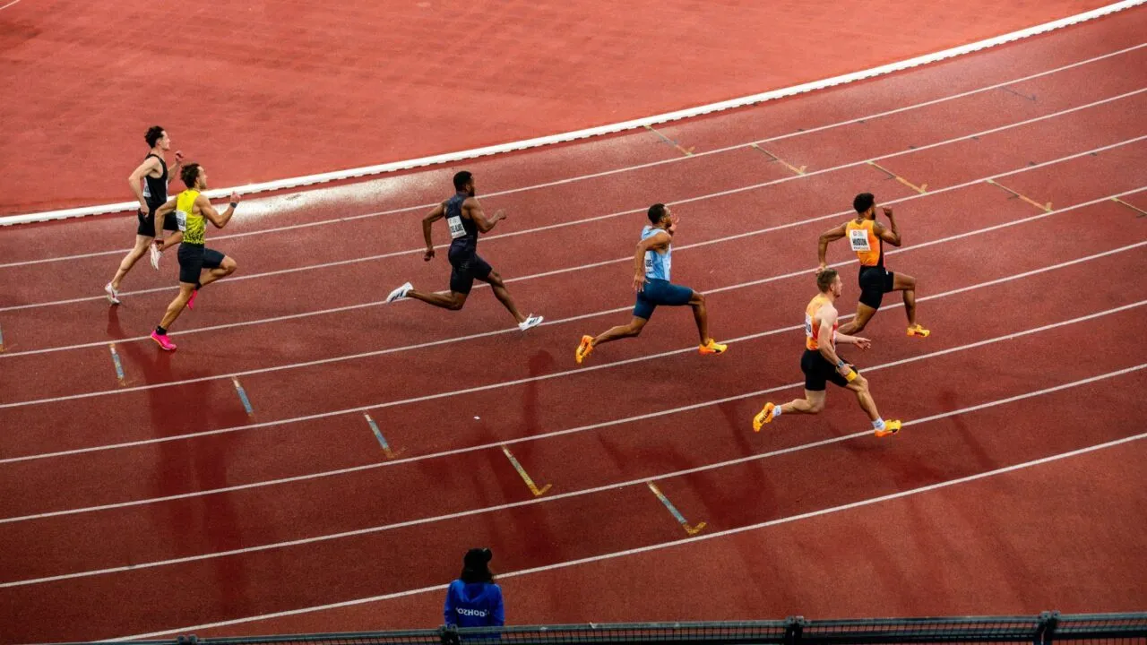Seks løbere sprinter på en buet del af en udendørs bane, hver i deres egen bane. En person i blåt står ved rækværket og observerer løbet. Banens overflade er rødbrun med hvide banemarkeringer.
