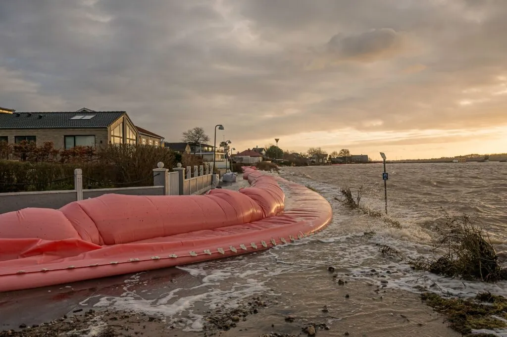 En stor lyserød oppustelig oversvømmelsesbarriere strækker sig langs kanten af et boligområde og holder det stigende stormvand tilbage, mens bølgerne bryder mod den under en overskyet, gylden himmel ved solnedgang.