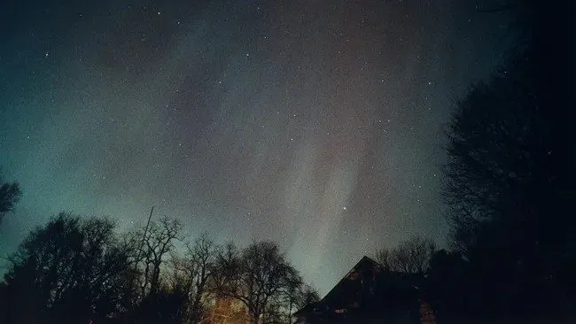 Nattehimmel med svage, nordlysagtige lysstriber, stjerner og silhuetter af nøgne træer og et hustag i forgrunden.