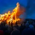 I skumringen samles en stor menneskemængde for at se på et kæmpe bål, der brænder lyst, mens høje flammer og tæt røg stiger op mod den dybe blå himmel.