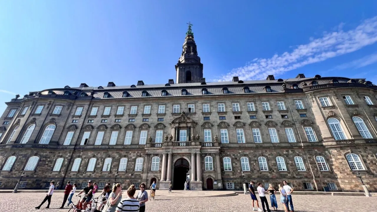 En stor historisk bygning med mange vinduer og et højt tårn i midten står under en klar blå himmel. Flere mennesker og cykler er samlet på den store brostensbelagte plads foran.