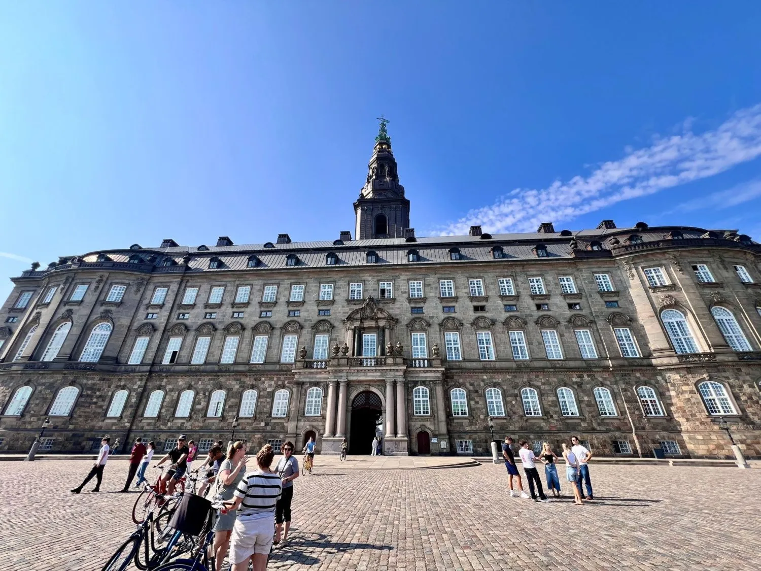 En stor historisk bygning med mange vinduer og et højt tårn i midten står under en klar blå himmel. Flere mennesker og cykler er samlet på den store brostensbelagte plads foran.
