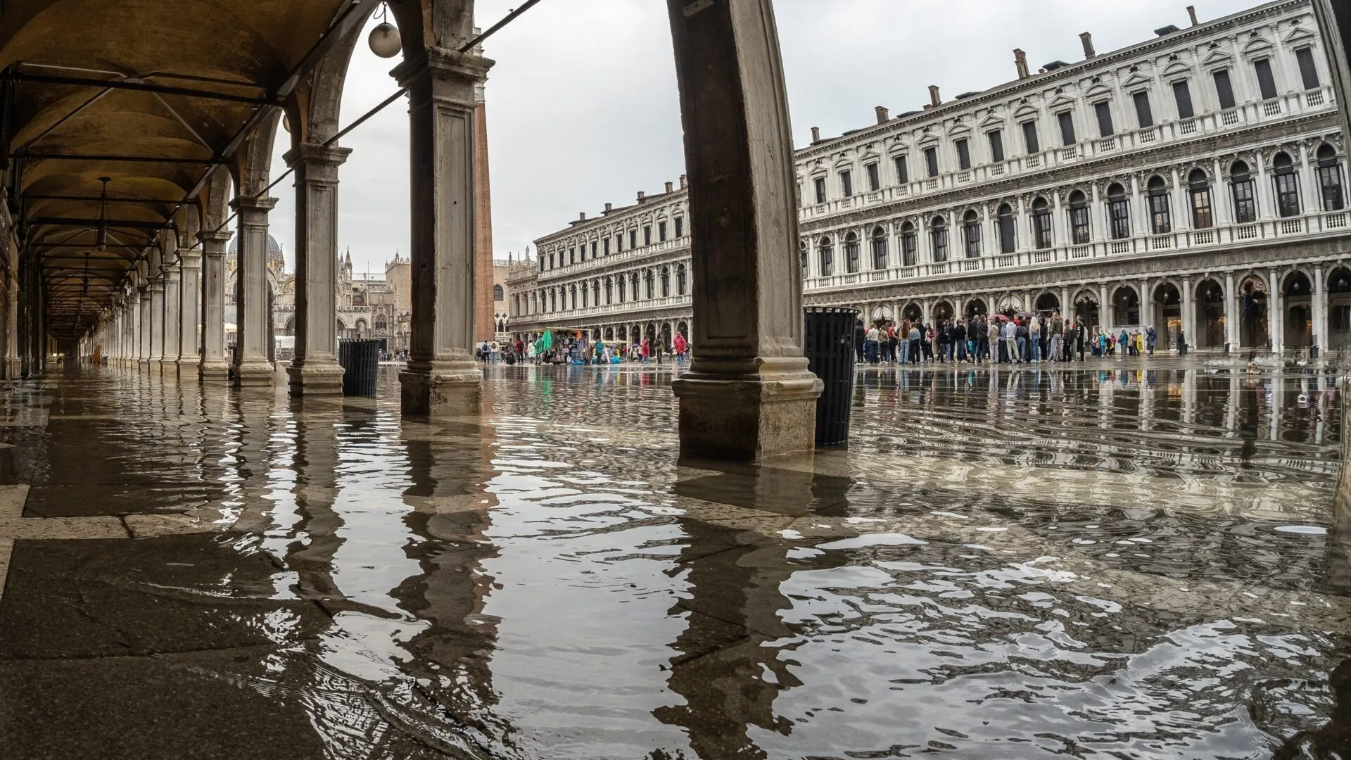 Et oversvømmet torv i Venedig, Italien, hvor vandet spejler den buede portik og de historiske bygninger. Der står mennesker i det fjerne, mens torvet er delvist oversvømmet.