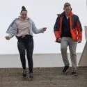 To personer, den ene i hættetrøje og den anden i rød jakke, går ned ad en udendørs betontrappe, mens de hver især holder fast i gelænderet. Himlen er overskyet.