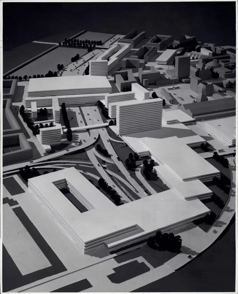 Et sort-hvidt foto af en arkitektonisk model, der viser et bybillede med mange moderne, rektangulære bygninger, veje og grønne områder, set oppefra i en vinkel.