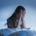 En person, der sidder op i sengen om natten med hovedet i hænderne og ser stresset eller oprevet ud. Værelset er svagt oplyst i blå nuancer, hvilket fremkalder en følelse af tristhed eller søvnløshed.