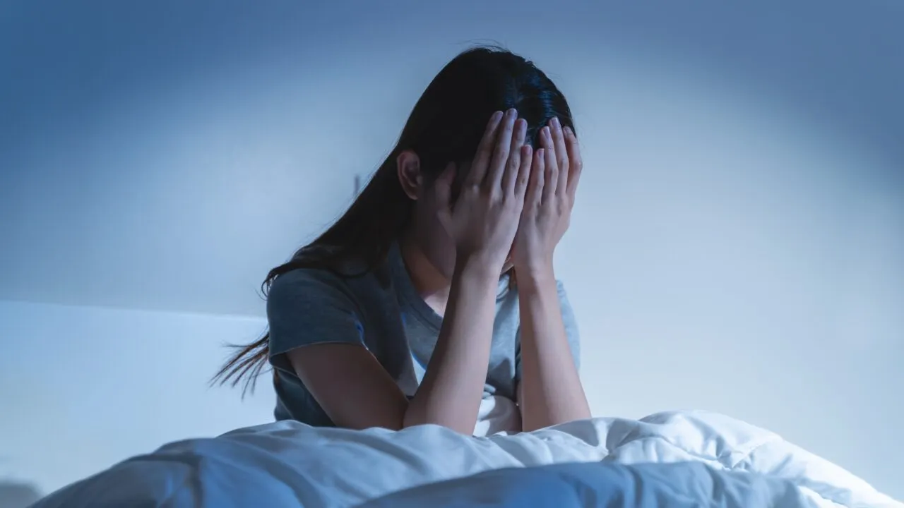 En person, der sidder op i sengen om natten med hovedet i hænderne og ser stresset eller oprevet ud. Værelset er svagt oplyst i blå nuancer, hvilket fremkalder en følelse af tristhed eller søvnløshed.