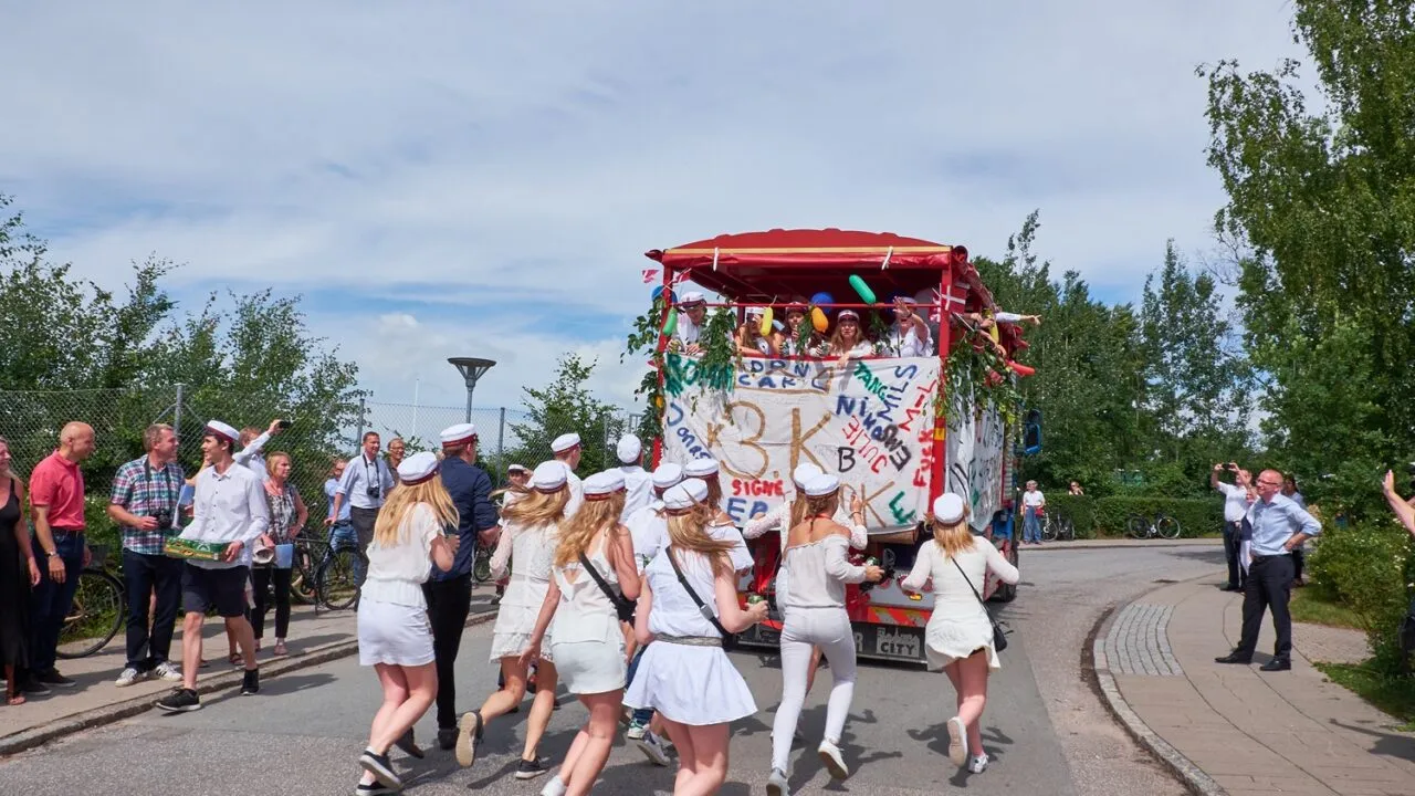 En gruppe studerende i hvide dragter og kasketter løber glædestrålende hen mod en pyntet lastbil, mens folk ser på fra siden af en gade, der er omgivet af træer og grønne områder under en delvis overskyet himmel.