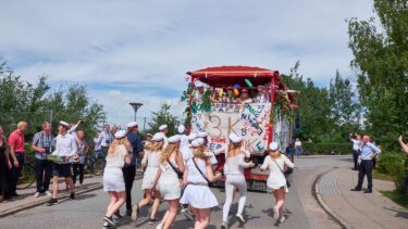 En gruppe studerende i hvide dragter og kasketter løber glædestrålende hen mod en pyntet lastbil, mens folk ser på fra siden af en gade, der er omgivet af træer og grønne områder under en delvis overskyet himmel.