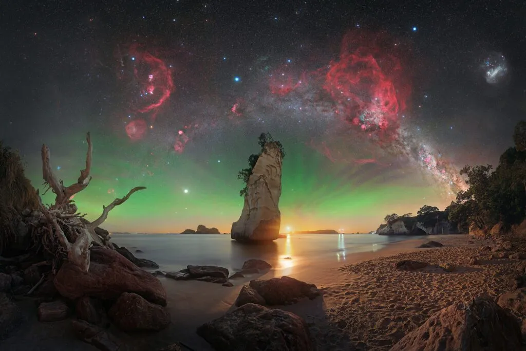 En dramatisk strandscene ved solnedgang med klippeskær, en høj klippestøtte, drivtømmer og roligt vand. Himlen lyser op af grøn nordlys og er fyldt med klare stjerner, røde tåger og Mælkevejen, der buer sig hen over himlen.
