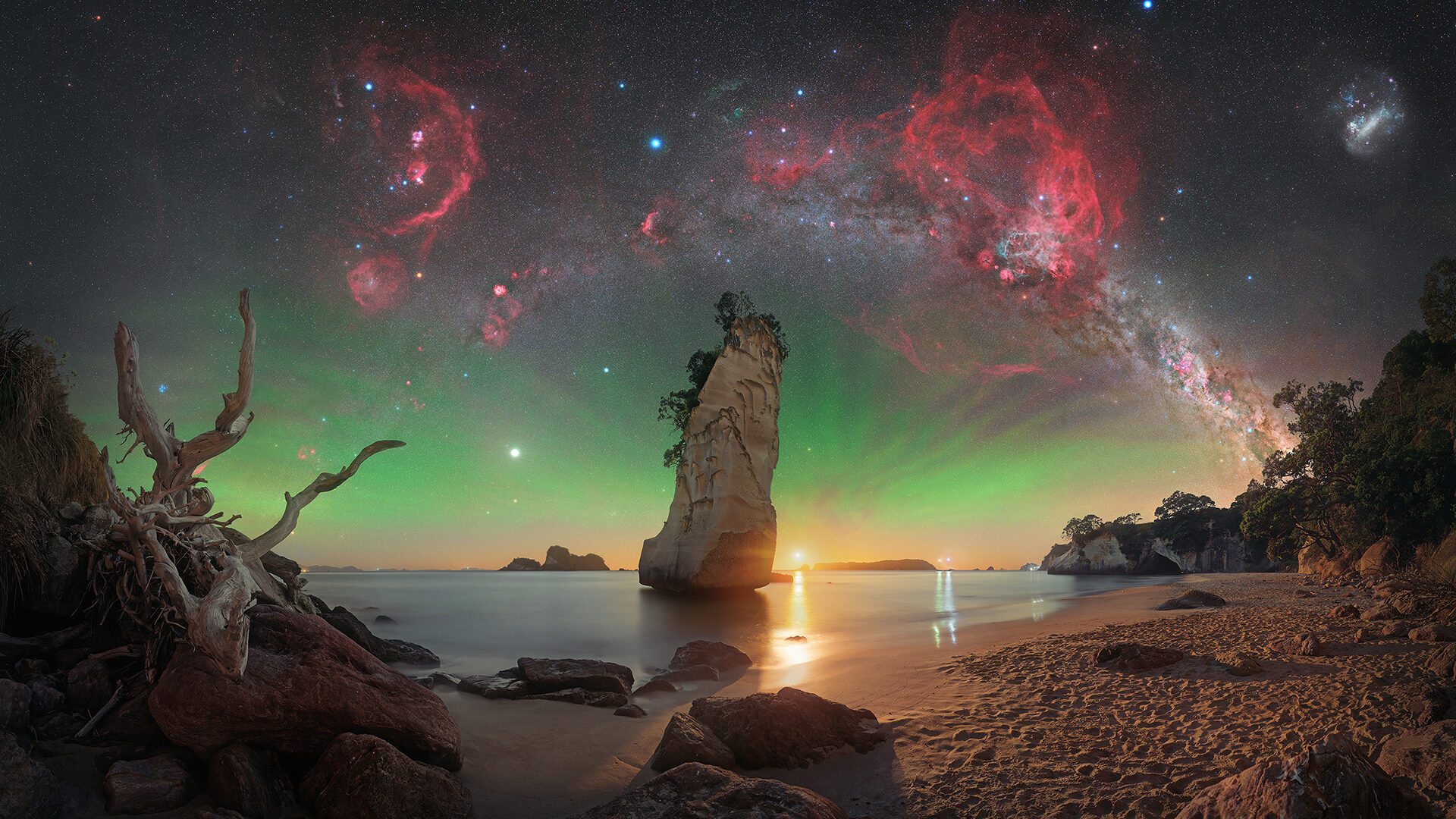 En dramatisk strandscene ved solnedgang med klippeskær, en høj klippestøtte, drivtømmer og roligt vand. Himlen lyser op af grøn nordlys og er fyldt med klare stjerner, røde tåger og Mælkevejen, der buer sig hen over himlen.