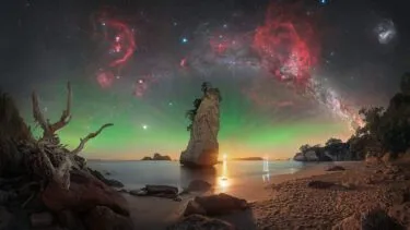 En dramatisk strandscene ved solnedgang med klippeskær, en høj klippestøtte, drivtømmer og roligt vand. Himlen lyser op af grøn nordlys og er fyldt med klare stjerner, røde tåger og Mælkevejen, der buer sig hen over himlen.