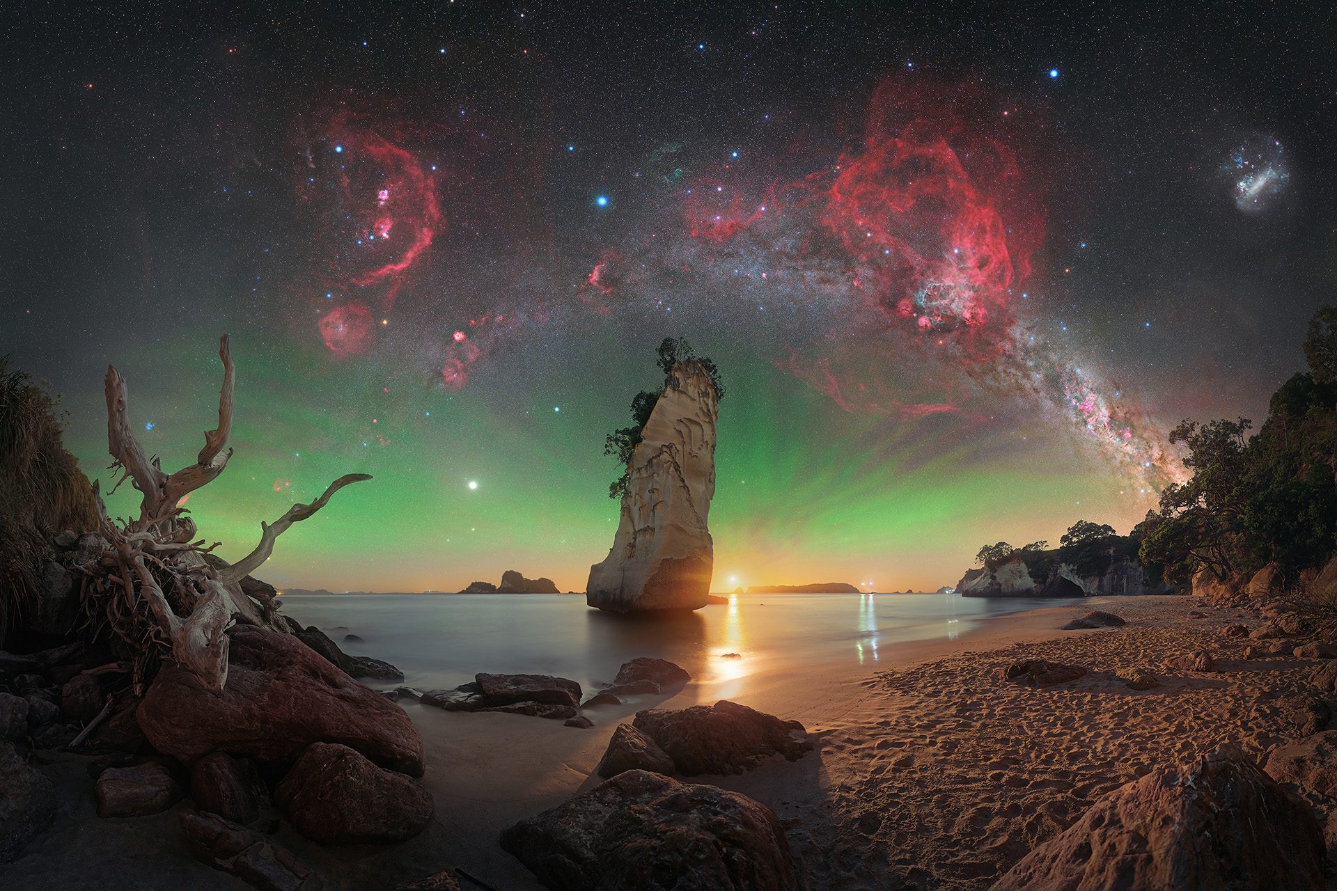 En dramatisk strandscene ved solnedgang med klippeskær, en høj klippestøtte, drivtømmer og roligt vand. Himlen lyser op af grøn nordlys og er fyldt med klare stjerner, røde tåger og Mælkevejen, der buer sig hen over himlen.