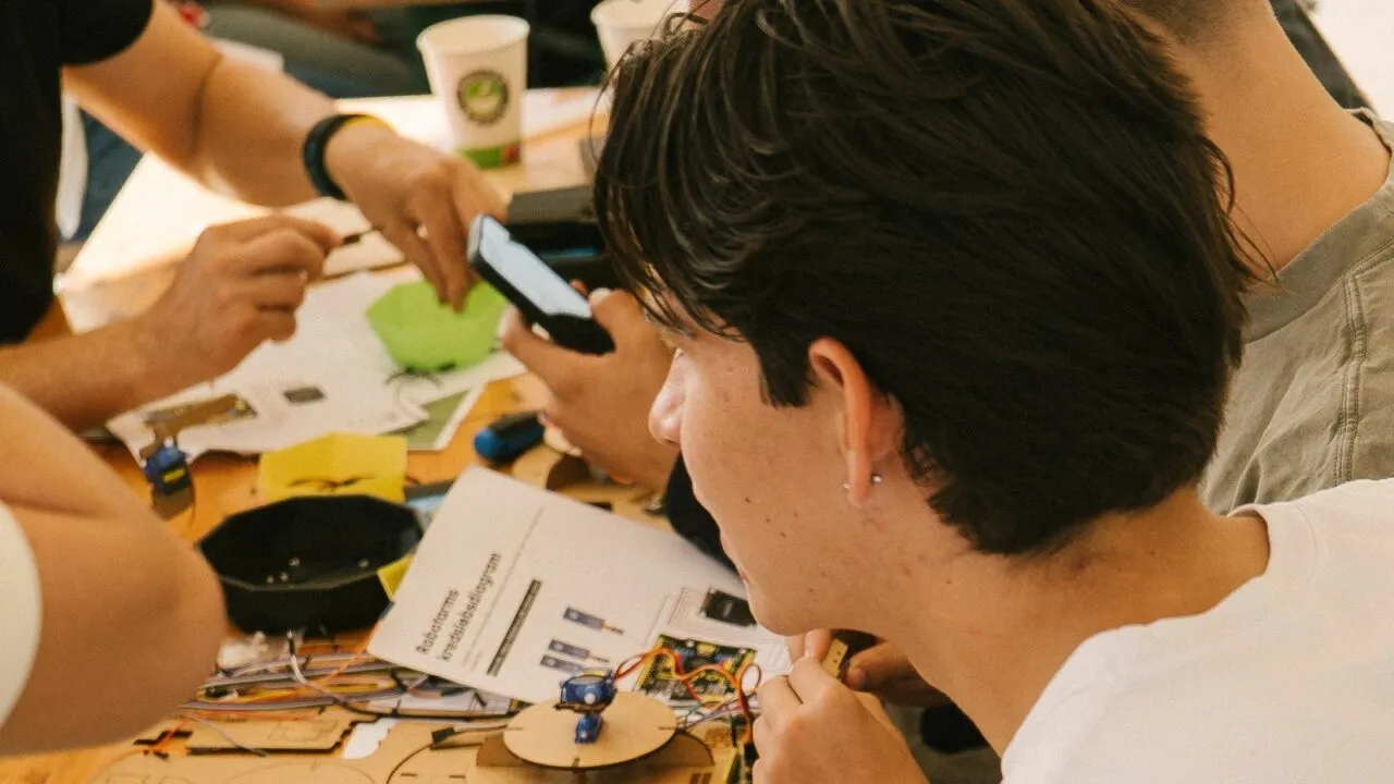En gruppe unge mennesker arbejder sammen om et praktisk elektronikprojekt ved et bord, hvor papirer, ledninger og komponenter er spredt ud. En person er fokuseret på projektet, mens en anden bruger en telefon.