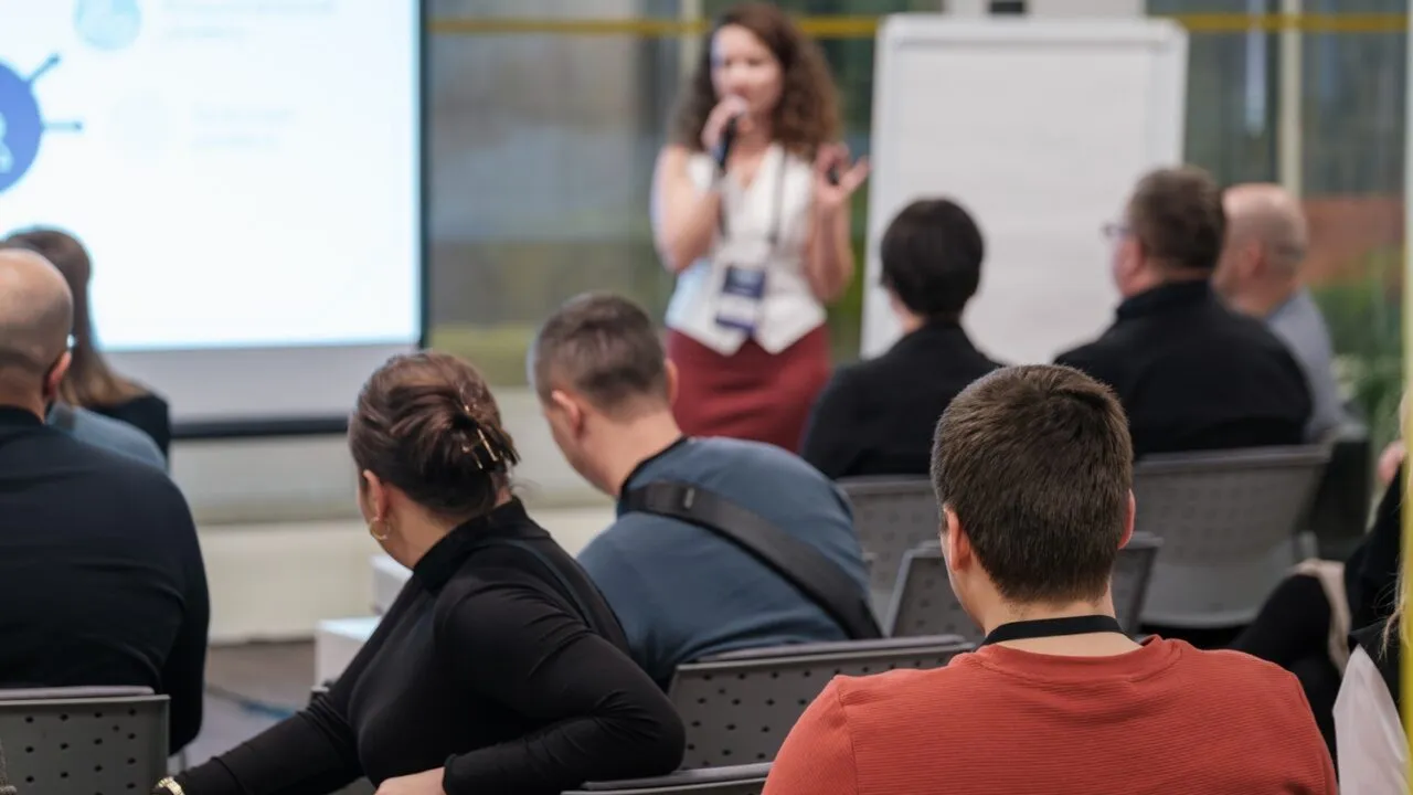 En kvinde taler i en mikrofon foran et siddende publikum ved et seminar eller en workshop, mens der i baggrunden ses en projektorskærm og et flipover-tavle.
