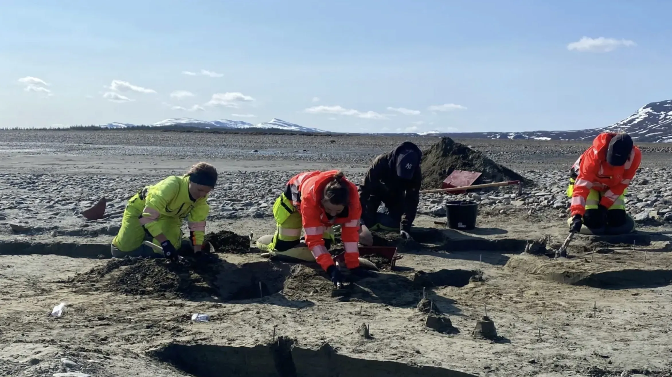 Tre personer i synligt arbejdstøj udfører gravearbejde på en sandet grund udendørs, med bjerge og sne i det fjerne i baggrunden under en klar himmel.