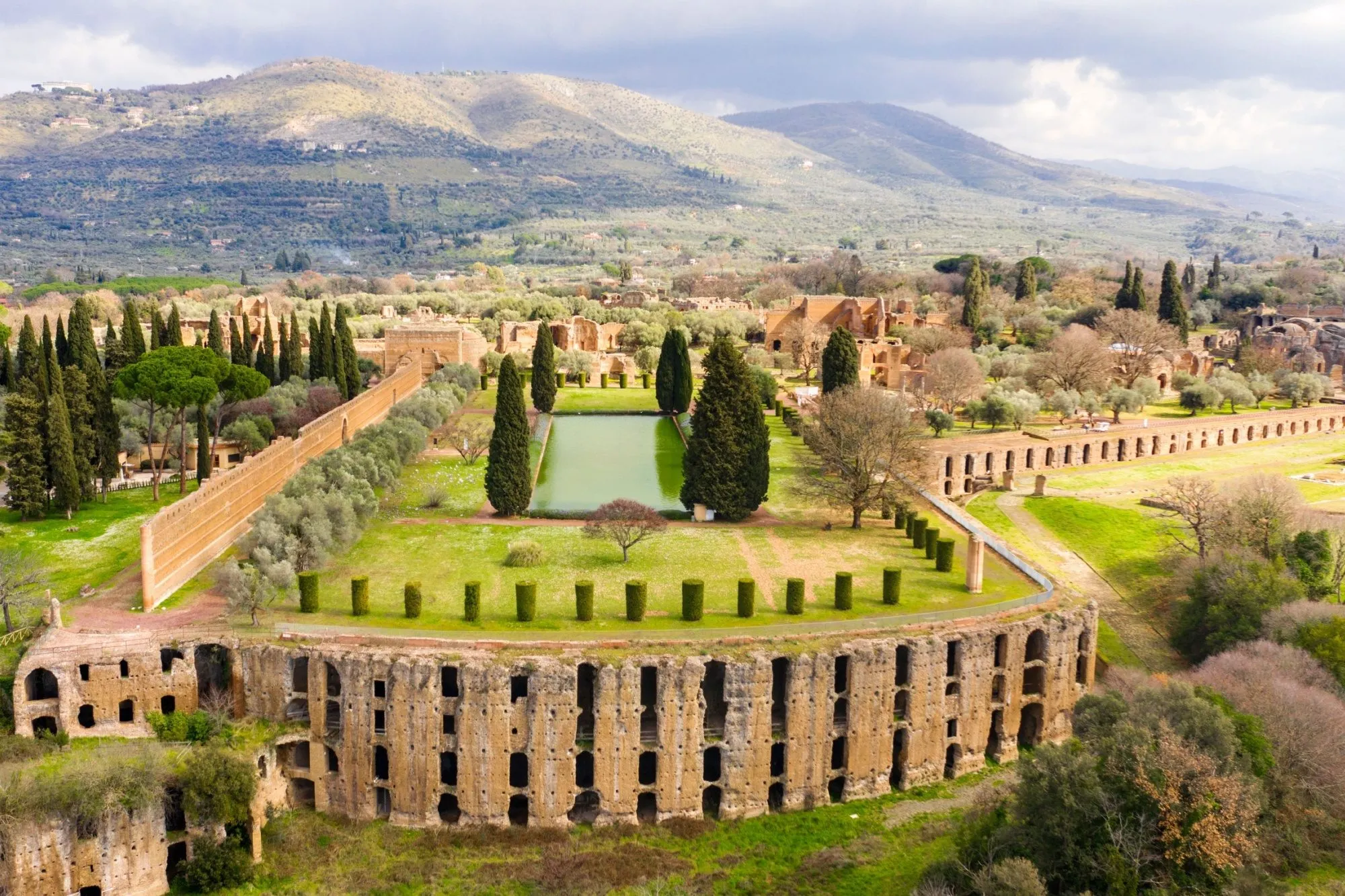 Luftfoto af de antikke ruiner af Hadrians Villa i Tivoli, Italien, der viser rektangulære bassiner, cypresser, stensøjler og de omgivende grønne bakker under en overskyet himmel.
