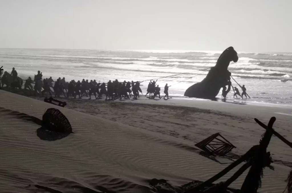 En gruppe mennesker trækker en stor, delvist nedgravet statue langs en strand ved solnedgang; bølgerne bruser tæt ved, og der ligger affald spredt ud over sandet.