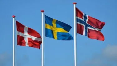 Tre flag på flagstænger mod en blå himmel: det rød-hvide danske flag, det blå-gule svenske flag og det rød-hvid-blå norske flag, som alle vajer i vinden.