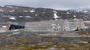 En moderne, kantet bygning med mange vinduer ligger i et klippefyldt tundralandskab med snepletter på jorden og bjerge i baggrunden under en overskyet himmel.