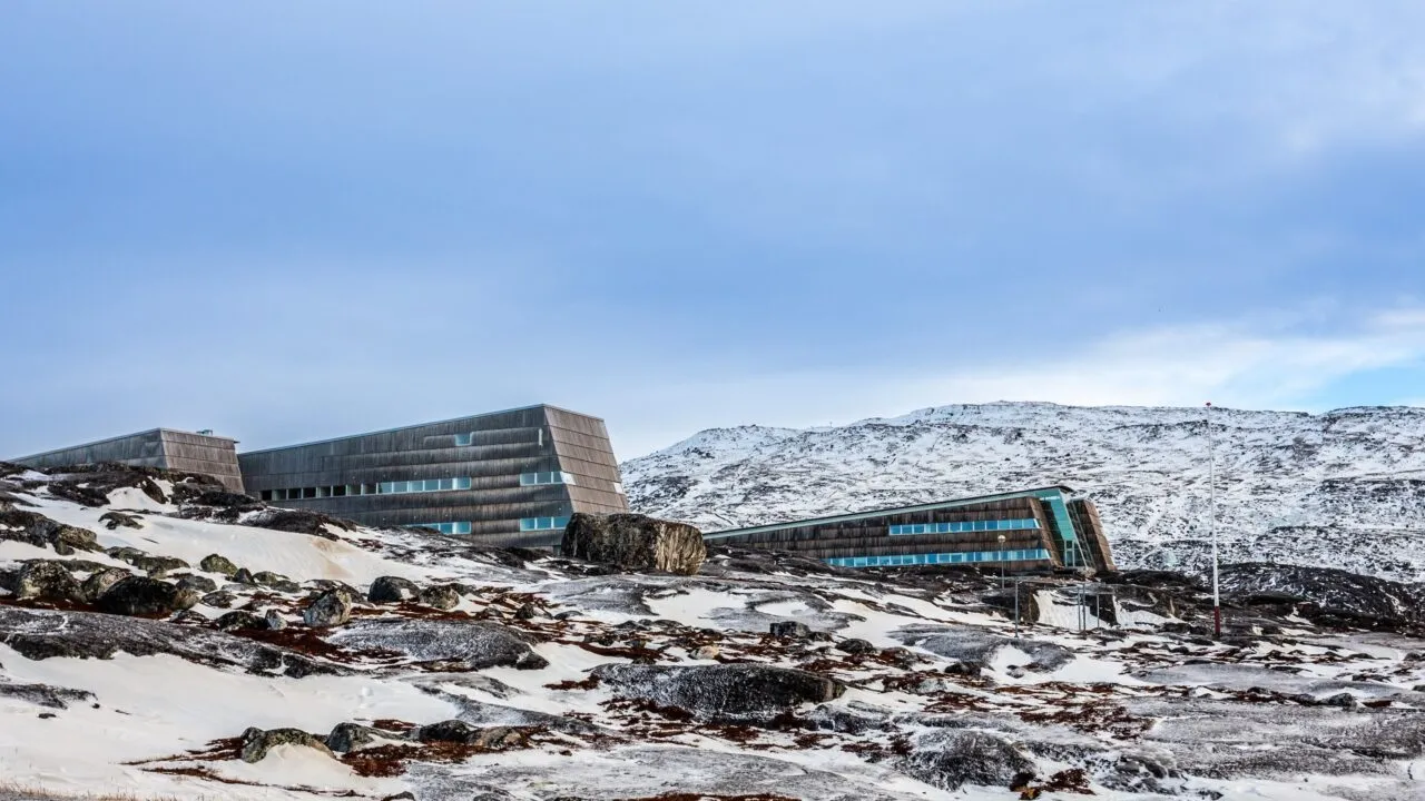 Moderne bygninger med grøntonede vinduer ligger på en klippefyldt, snedækket bjergskråning under en overskyet himmel og smelter sammen med det barske arktiske landskab.