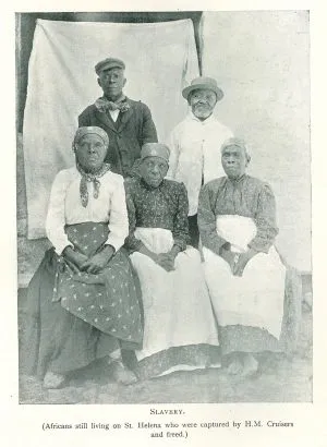 Fem ældre voksne, hvoraf tre sidder og to står, poserer foran et lysskærm. Billedteksten angiver, at der er tale om afrikanere på St. Helena, som blev befriet fra slaveri af H.M. krydsere. Historisk sort-hvidt fotografi.