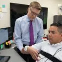 En læge undersøger en mand, der sidder med en hvid anordning om håndleddet og på hovedet, mens der står medicinsk udstyr og computerudstyr på skrivebordene i et kliniklokale.