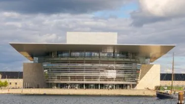 En moderne bygning ved vandet med et fladt, udhængende tag og en glasfacade ligger ved vandet under en delvis overskyet himmel.
