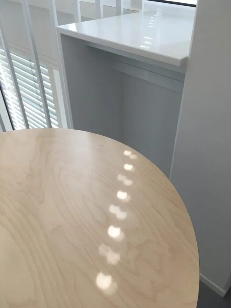 Et rundt bord af lyst træ med solpletter står tæt ved et hvidt gelænder og en lille, hvid hylde ved et vindue med persienner.