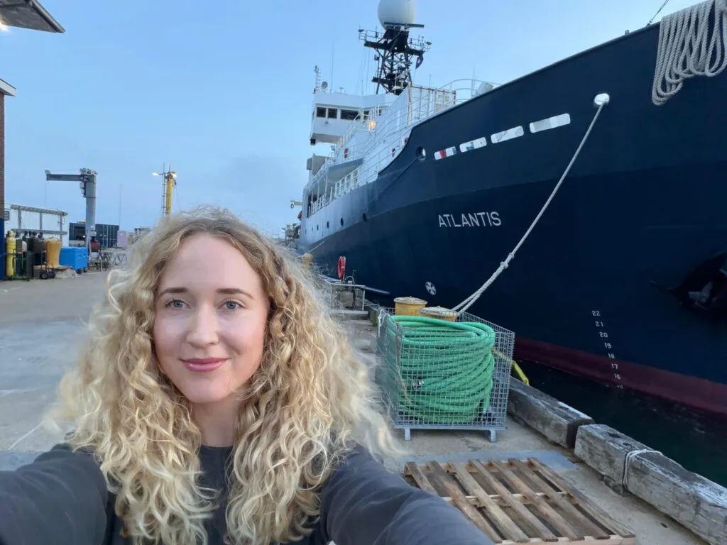 En kvinde med krøllet blondt hår tager et selfie på en kaj foran et stort skib ved navn Atlantis. På kajen ved siden af hende kan man se sammenrullede grønne slanger og udstyr.
