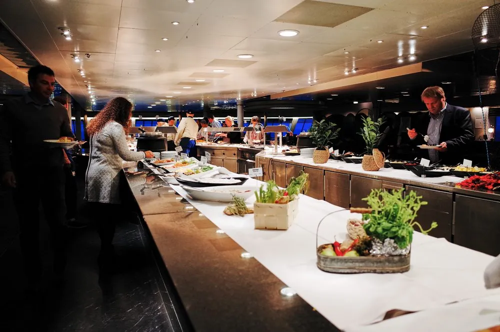 En buffet med forskellige retter udstillet på en lang disk, hvor flere personer tager for sig af maden under kraftig indendørsbelysning. Stemningen virker livlig og moderne.