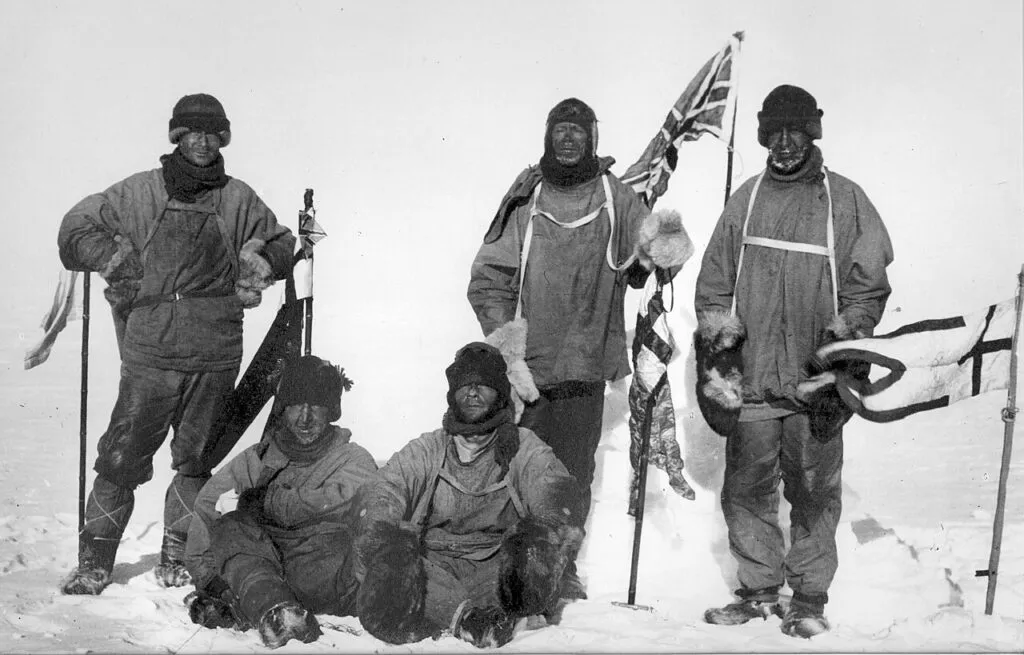 Fem opdagelsesrejsende i kraftigt vintertøj poserer i sneen; to står med flag, mens tre sidder ned. Scenen fremstår barsk og blæsende, med sne på jorden og udstyr synligt ved siden af dem.
