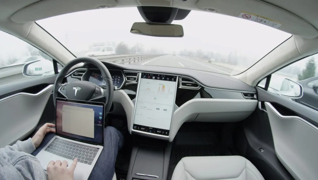 Et billede fra kabinen i en Tesla-bil, der kører på motorvejen i autopilot-tilstand. En person på førersædet bruger en bærbar computer med hænderne væk fra rattet, og den store berøringsskærm viser kørselsdata.