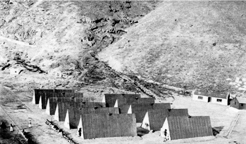 Et sort-hvidt foto viser rækker af hytter med trekantede tage, der ligger spredt i et goldt, kuperet landskab. Der ses et par små figurer rundt om bygningerne.