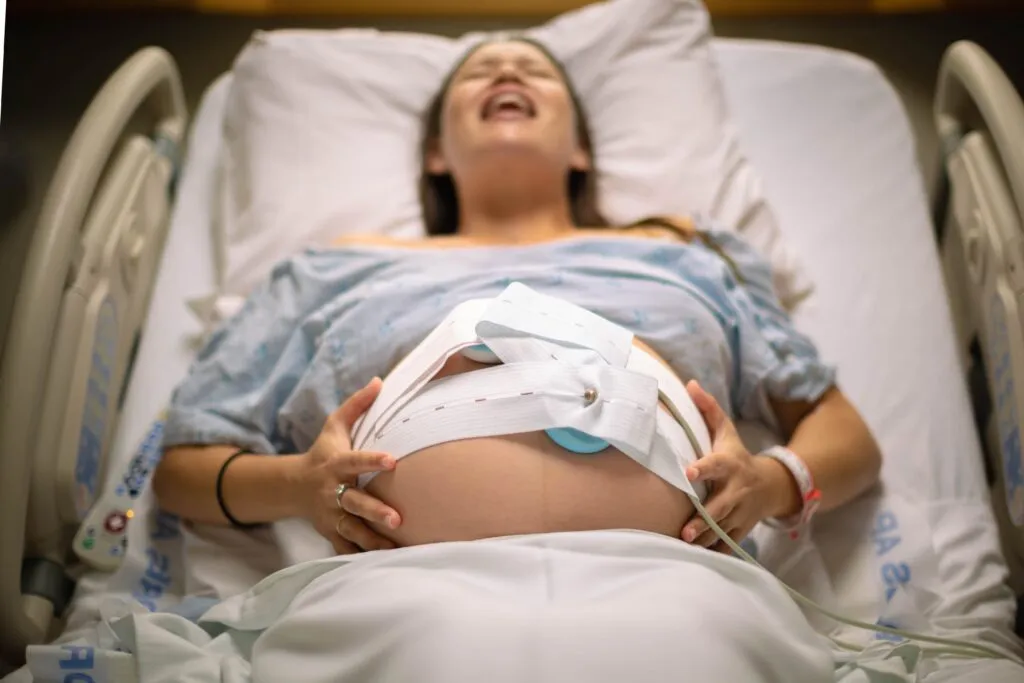 En gravid kvinde i en hospitalsseng, iført en hospitalskåbe, holder hånden mod maven, hvor der er fastgjort monitorer; hun ser ud til at være i fødsel og oplever kraftige veer.
