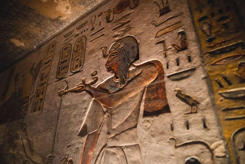 Et nærbillede af en gammel egyptisk vægudskæring, der viser en farao i profil, iført en hovedbeklædning og med en genstand i hånden, omgivet af hieroglyffer og dyresymboler. Væggen er oplyst af et varmt, gyldent lys.