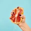 En hånd med mørkerød neglelak presser en halv grapefrugt, hvor saften drypper ned, på en ensfarvet lyseblå baggrund.