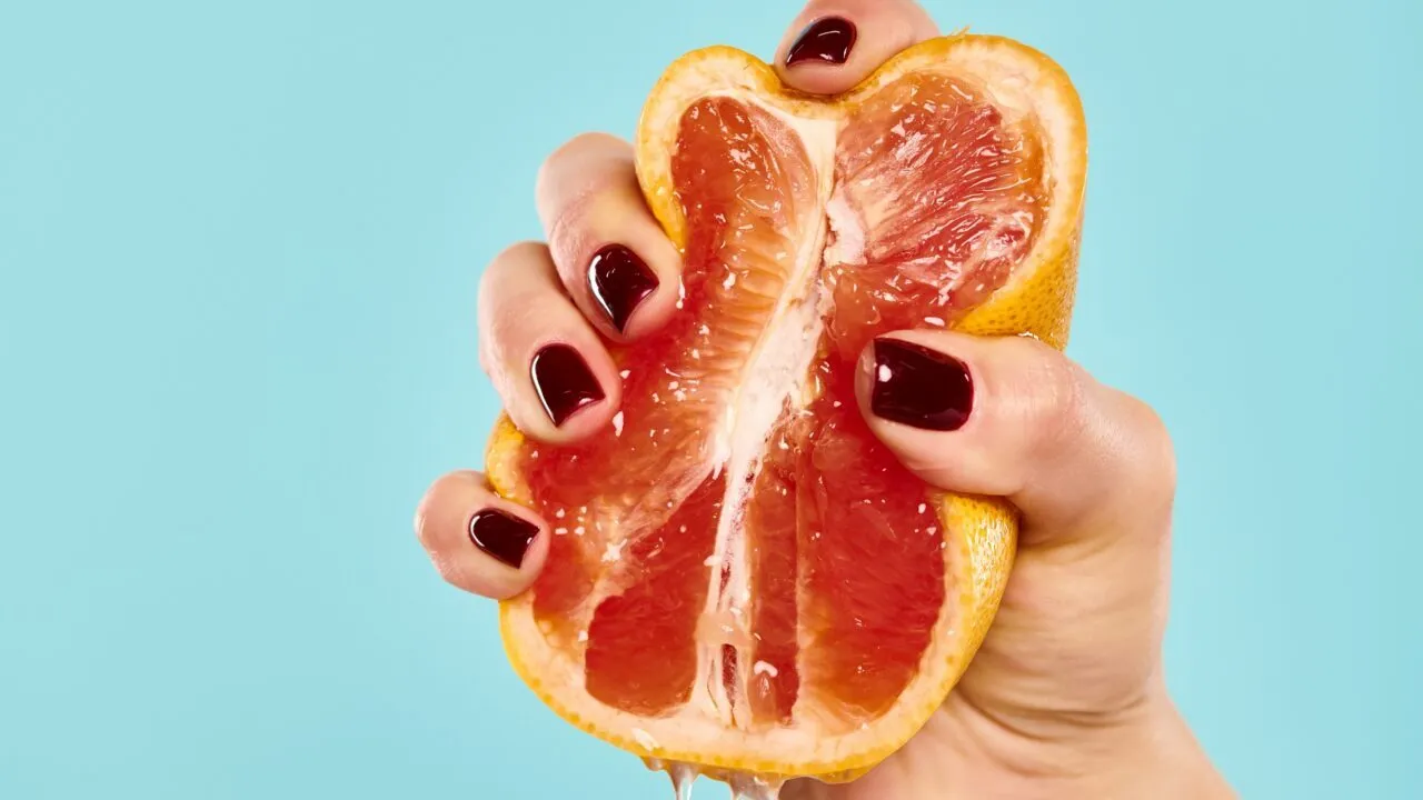 En hånd med mørkerød neglelak presser en halv grapefrugt, hvor saften drypper ned, på en ensfarvet lyseblå baggrund.