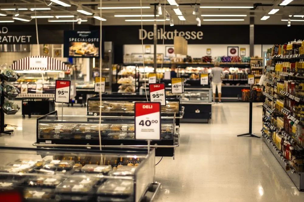 En delikatesseafdeling i et supermarked med køledisker, hylder med varer og røde prisskilte. Området er godt oplyst, og på bagvæggen står der »Delikatesse«, mens der står en person i baggrunden.