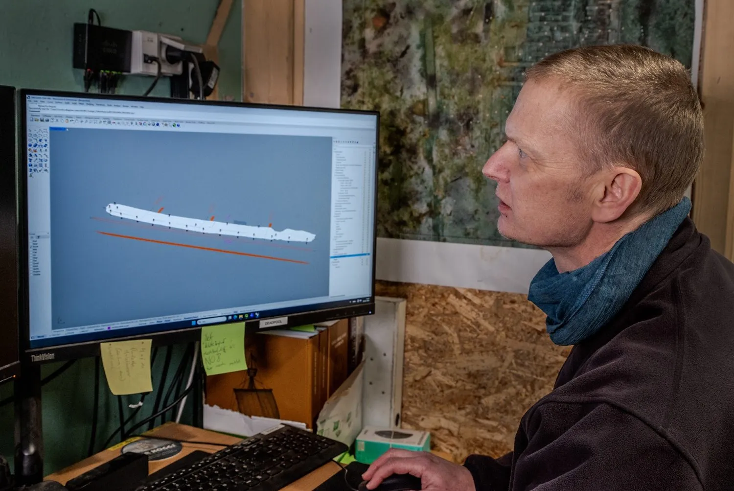 En person betragter en 3D-model af et langt objekt, muligvis et skibsvrag, på en computerskærm i et arbejdsrum ved hjælp af en mus.