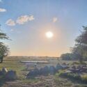 Et malerisk udendørsområde ved solnedgang med en strålende sol, grønt græs, store klipper, picnicborde og træer, der indrammer udsigten. Himlen er stort set klar med nogle få små skyer.
