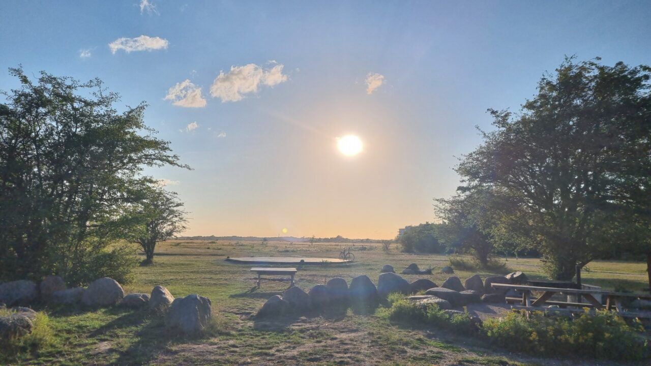 Et malerisk udendørsområde ved solnedgang med en strålende sol, grønt græs, store klipper, picnicborde og træer, der indrammer udsigten. Himlen er stort set klar med nogle få små skyer.
