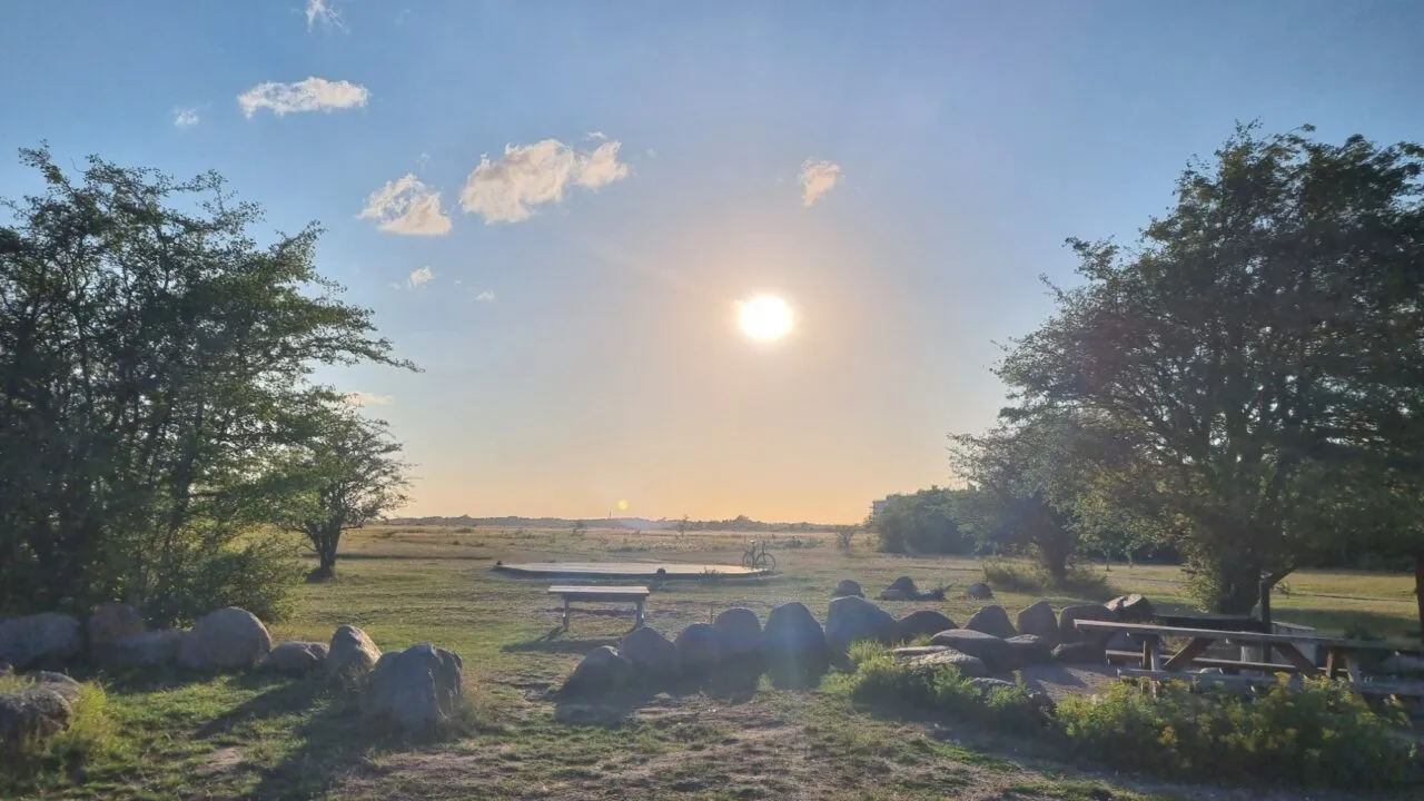 Et malerisk udendørsområde ved solnedgang med en strålende sol, grønt græs, store klipper, picnicborde og træer, der indrammer udsigten. Himlen er stort set klar med nogle få små skyer.