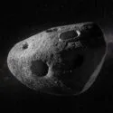 En grå, klippefyldt asteroide med adskillige store kratere svæver i det mørke rum. I baggrunden ses spredte stjerner og et svagt, tåget bånd, der ligner Mælkevejen.